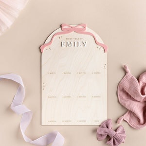 May include: A wooden growth chart with a pink bow and the text "First Year of Emily" on the top. The chart has spaces for each month of the baby's first year.