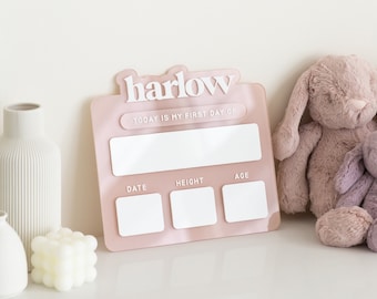 May include: Pink acrylic growth chart with the name "harlow" in white lettering. The chart has spaces to fill in the date, height, and age.  "Today is my first day of" is written above the blank space.