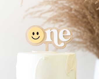 May include: A wooden cake topper with the word "one" in white letters and a yellow smiley face. The topper is on a white cake.