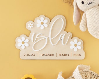 May include: A wooden sign with the name "Isla" written in cursive script, surrounded by white daisy flowers. The sign also includes the date "2.15.23", time "10:32am", weight "8.5lbs", and length "20in".