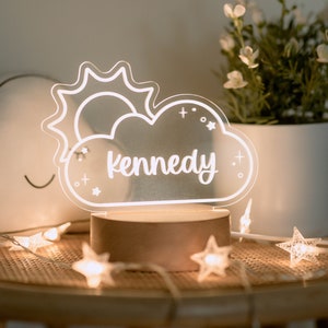 May include: A personalized night light featuring a cloud and sun design with the name "Kennedy" in white script. The light sits on a wooden base and is surrounded by star-shaped string lights. The Harper & Lo logo is in the top left corner.