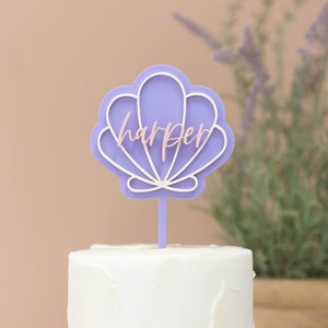 May include: A lavender seashell-shaped cake topper with the name "harper" in a light pink script. The topper is on a white cake, with a neutral background and the text "harper & lo" in the top left corner.