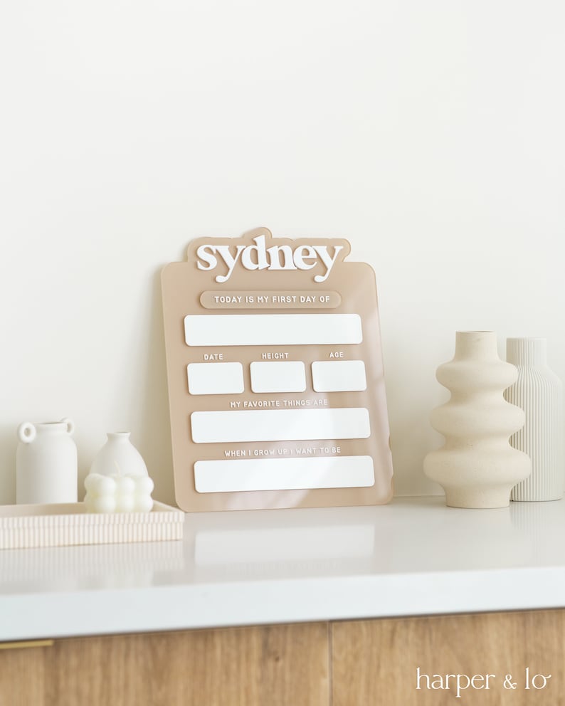 May include: A beige acrylic growth chart with the name "sydney" at the top. The chart has spaces to fill in the date, height, age, favorite things, and what the child wants to be when they grow up.