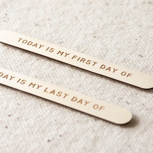 Pu&ograve; includere: Due bastoncini di legno con il testo "TODAY IS MY FIRST DAY OF" e "DAY IS MY LAST DAY OF" inciso su di essi. I bastoncini sono su uno sfondo di tessuto bianco.