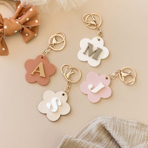 May include: Four flower-shaped keychains with gold rings and letters A, J, M, and S in different colors. The keychains are on a beige surface.