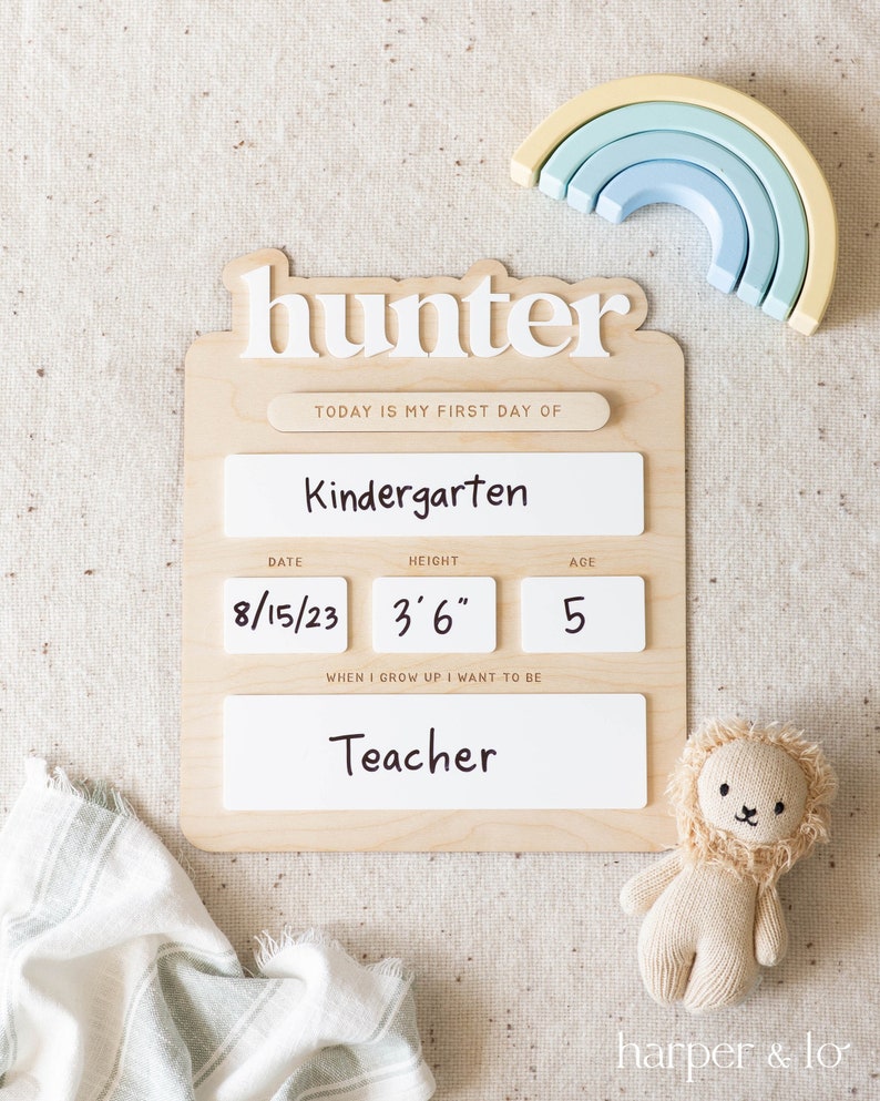 May include: A wooden sign with the name "hunter" written in white letters. The sign has a white background with black text that says "Today is my first day of Kindergarten". There are three white boxes with black text that say "Date", "Height", and "Age". The boxes have the following information filled in: 8/15/23, 3'6", and 5. The sign also has a white box with black text that says "When I grow up I want to be" and a white box with black text that says "Teacher".