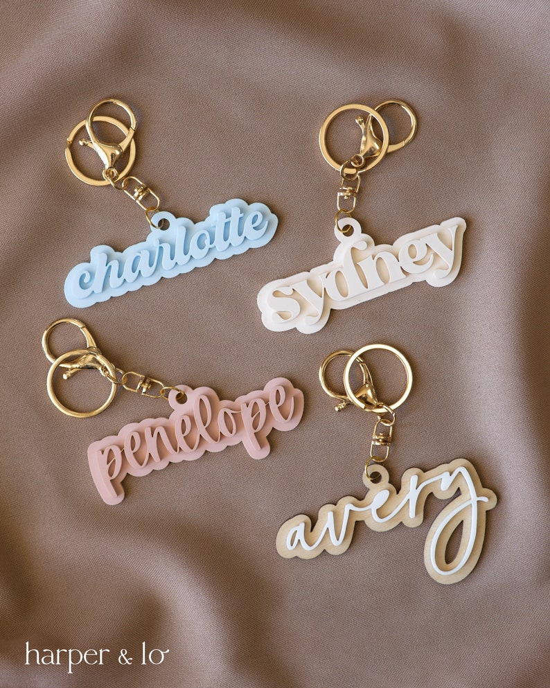 May include: Four acrylic keychains with gold key rings. The keychains are in the shape of names: Charlotte in light blue, Sydney in white, Penelope in pink, and Avery in beige with white lettering.