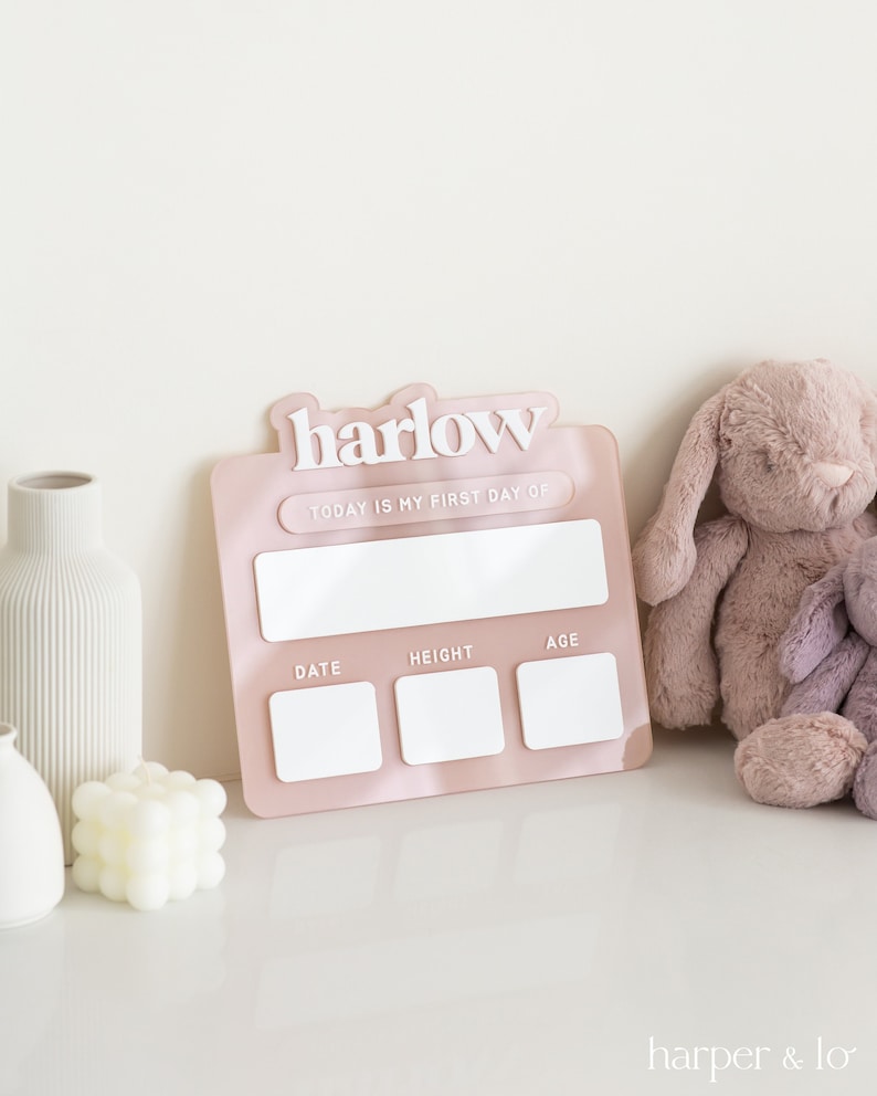 May include: Pink acrylic growth chart with the name "harlow" in white lettering. The chart has spaces to record the date, height, and age. The chart is on a white surface with a white vase and a stuffed bunny.