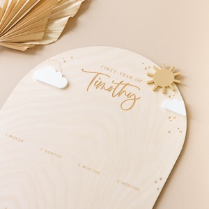 May include: A wooden growth chart with a light brown background and a white arch. The arch has the text "FIRST YEAR OF Timothy" in a cursive font. There are small white clouds and a yellow sun on the arch. The chart has numbers 1 through 7 for each month of the baby's first year.