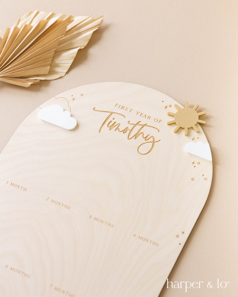 May include: A wooden growth chart with a light brown background and a white arch. The arch has the text "FIRST YEAR OF Timothy" in a cursive font. There are small white clouds and a yellow sun on the arch. The chart has numbers 1 through 7 for each month of the baby's first year.
