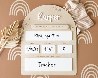 May include: A wooden first day of school sign with the name "Harper" in white script. The sign includes fields for "Kindergarten", date, height (107 cm), age (5), and "Teacher". A tan and white striped shirt is next to the sign.