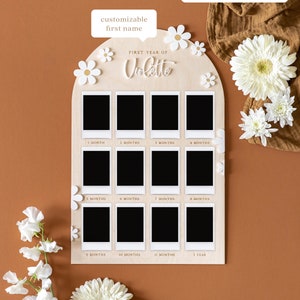 May include: A wooden photo display with a white arch shape and 12 photo slots. The display is decorated with white flowers and the text "FIRST YEAR OF Violette".