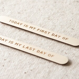 May include: Two wooden sticks with the text "TODAY IS MY FIRST DAY OF" and "DAY IS MY LAST DAY OF" engraved on them. The sticks are on a white fabric background.