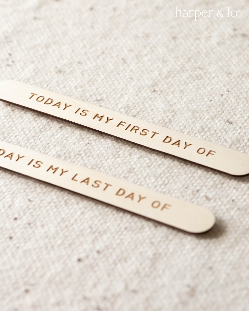 May include: Two wooden sticks with the text "TODAY IS MY FIRST DAY OF" and "DAY IS MY LAST DAY OF" engraved on them. The sticks are on a white fabric background.