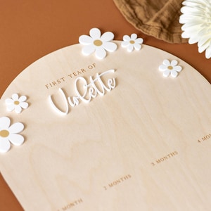May include: A wooden growth chart with a white arch and the words "FIRST YEAR OF" and "Volatte" in gold lettering. The chart has spaces for each month of the baby's first year, with white flower decorations.