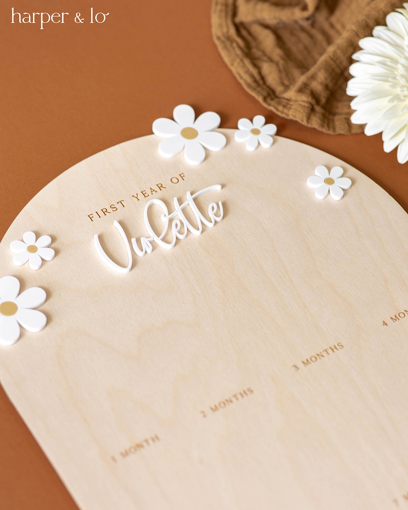 May include: A wooden growth chart with a white arch and the words "FIRST YEAR OF" and "Volatte" in gold lettering. The chart has spaces for each month of the baby's first year, with white flower decorations.