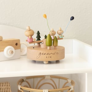 May include: Wooden music box with a pink, blue, and yellow color scheme. The music box features a seesaw with two figures and two trees. The music box is engraved with the name "Genevieve Kim".
