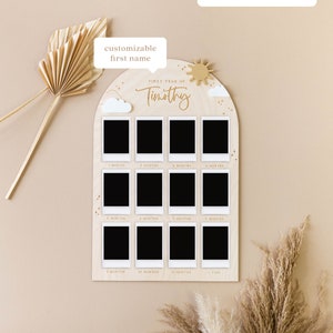May include: A wooden photo display with a light brown background and a white arch. The arch has a sun and clouds on it. The arch has 12 photo slots for pictures of a baby's first year. The text "FIRST YEAR OF" and "Timothy" are written in gold on the arch. The text "customizable first name" is written in a speech bubble above the arch. The text "polaroids/printed photos not included." is written in a speech bubble above the arch.
