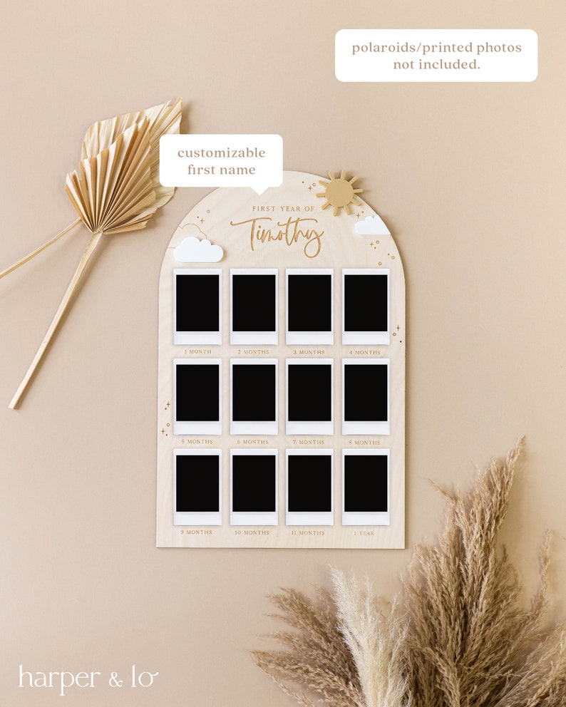 May include: A wooden photo display with a light brown background and a white arch. The arch has a sun and clouds on it. The arch has 12 photo slots for pictures of a baby's first year. The text "FIRST YEAR OF" and "Timothy" are written in gold on the arch. The text "customizable first name" is written in a speech bubble above the arch. The text "polaroids/printed photos not included." is written in a speech bubble above the arch.