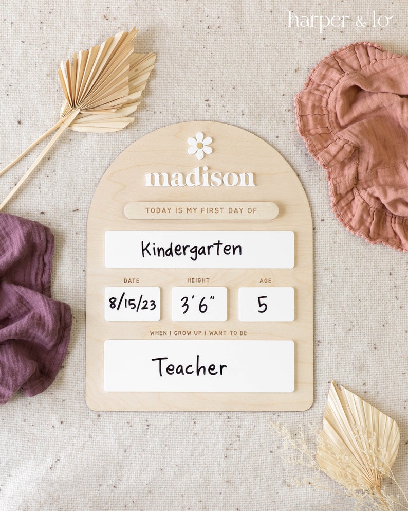 Pu&ograve; includere: Un cartello di legno con una margherita bianca e il nome "madison" scritto in lettere bianche. Il cartello dice "Oggi &egrave; il mio primo giorno di scuola materna" e include la data "8/15/23", l'altezza "3'6"