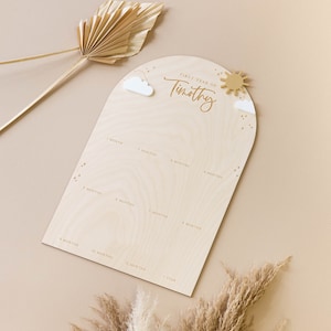 May include: A wooden growth chart with a light brown background and a white arch at the top. The arch has a sun and clouds with a white star. The text "First Year of Timothy" is written in a cursive font. The chart has 12 months and 1 year marked on it.