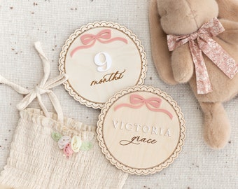 May include: Two wooden baby milestone discs with pink bows. One disc says "9 months" and the other says "Victoria Grace".
