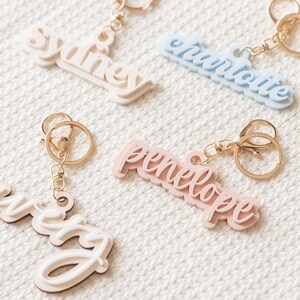 May include: Four personalized keychains with names Sydney, Charlotte, Penelope, and Avery. The keychains are made of layered acrylic in different colors: white, light blue, pink, and cream. Each keychain has a gold metal ring and a gold metal clasp.