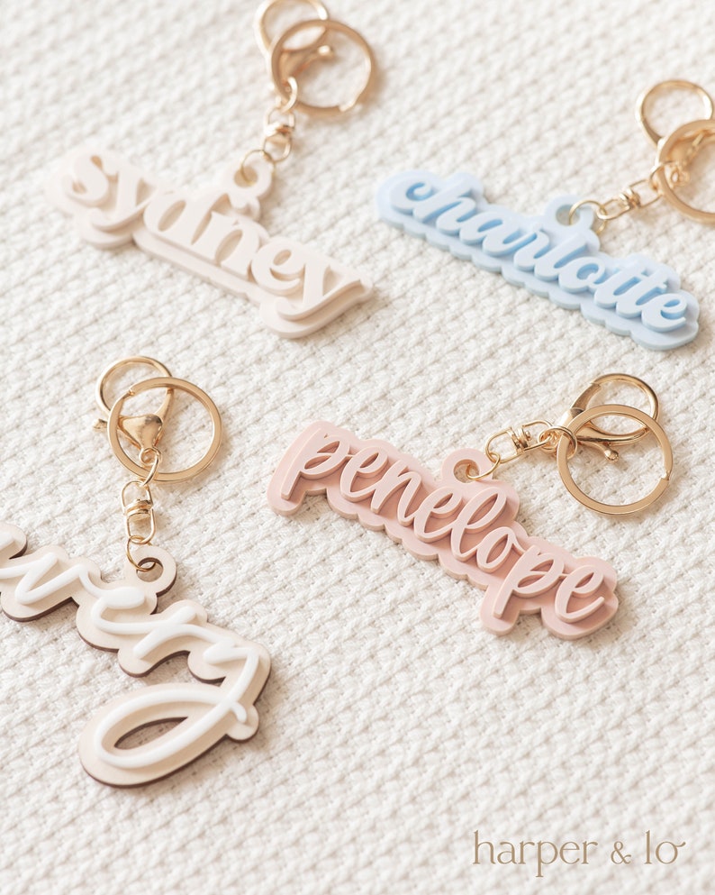 May include: Four personalized keychains with names Sydney, Charlotte, Penelope, and Avery. The keychains are made of layered acrylic in different colors: white, light blue, pink, and cream. Each keychain has a gold metal ring and a gold metal clasp.