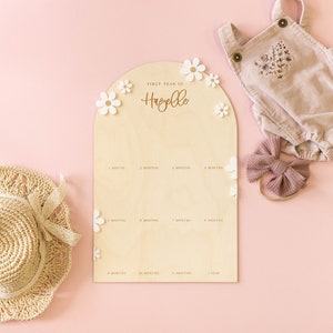 May include: A wooden growth chart with a light brown background and white flowers. The chart is shaped like an arch and has the text "FIRST YEAR OF Hazelle" at the top. The chart has 12 spaces for marking a child's growth, each labeled with a month.