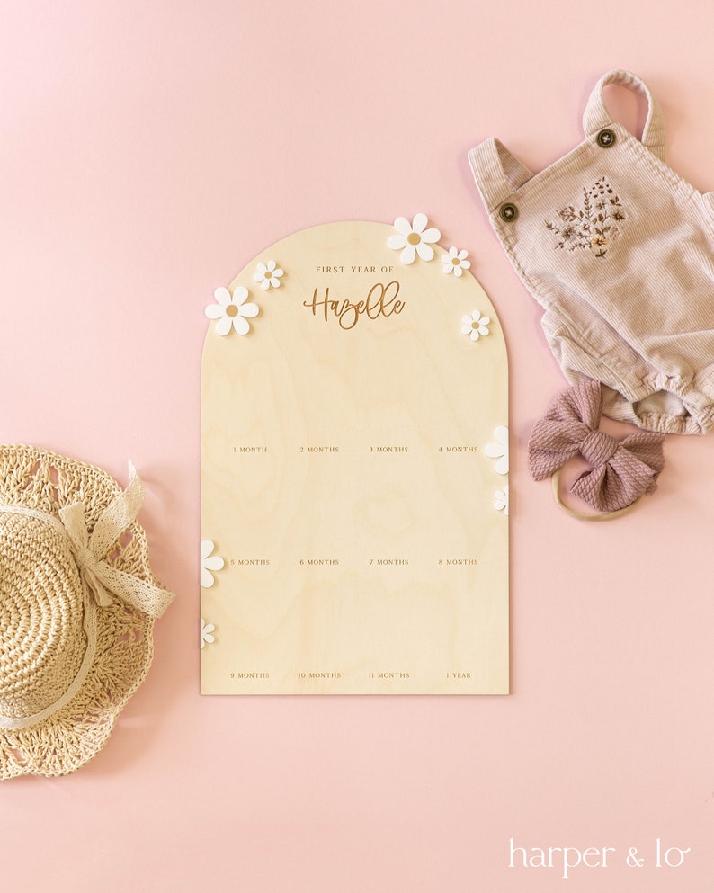 May include: A wooden growth chart with a light brown background and white flowers. The chart is shaped like an arch and has the text "FIRST YEAR OF Hazelle" at the top. The chart has 12 spaces for marking a child's growth, each labeled with a month.