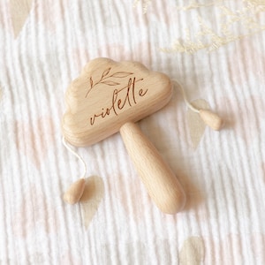 May include: A wooden baby rattle in the shape of a cloud, engraved with the name "violette" and a leaf design. The rattle has a handle and two wooden beads attached by strings. The background is a white and pink patterned fabric.