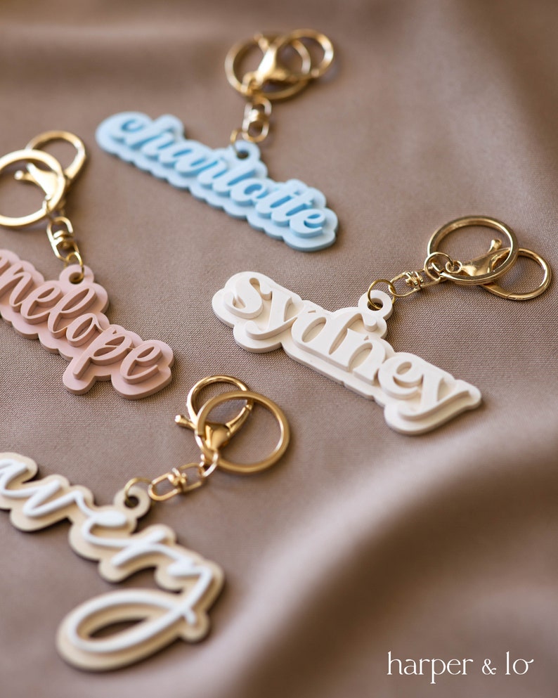 May include: Four personalized keychains with gold rings. The names Charlotte, Sydney, Melody, and Kristy are written in a layered, 3D style. The keychains are on a brown fabric background.