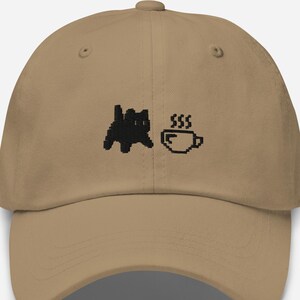 May include: A beige baseball cap with a black pixelated cat and a pixelated coffee cup with steam. The cap has a curved peak and an adjustable strap. The design is on the front of the cap.