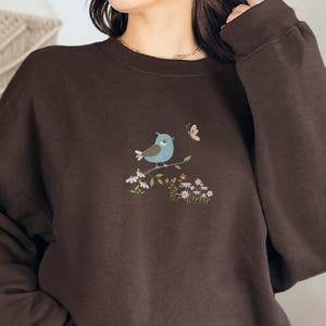 May include: A dark brown sweatshirt featuring an embroidered design of a blue bird perched on a branch with flowers and a butterfly. The bird has a light blue body and is facing left. The butterfly is pink. The sweatshirt has long sleeves.