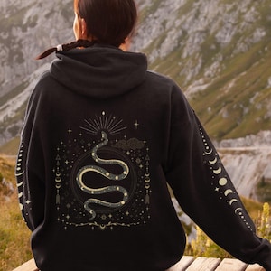 May include: Black hooded sweatshirt with a gold snake and celestial design on the back. The design features a snake wrapped around a circle with stars, moons, and other celestial symbols.