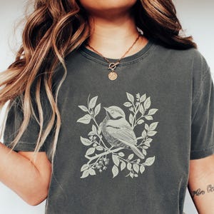 Op de afbeelding: Donkergrijs T-shirt met een gedetailleerde illustratie van een vogel op een tak met bladeren. De vogel en het gebladerte zijn in een lichtere tint, wat contrasteert met de achtergrond van het shirt. Het shirt heeft een klassieke ronde hals en korte mouwen.