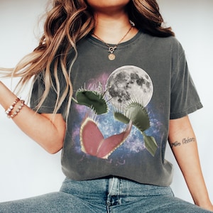 May include: Dark grey t-shirt featuring a Venus flytrap design with a full moon and a galaxy background. The graphic includes a pink and green flytrap, with a detailed moon and a colorful nebula.