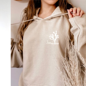 May include: A beige hoodie with a white Joshua Tree graphic on the front. The hoodie has a drawstring hood and a kangaroo pocket.