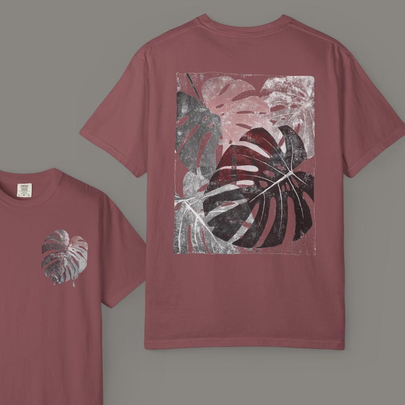 May include: A maroon t-shirt featuring a large, distressed print of monstera leaves in shades of gray, black, and burgundy on the back. A smaller version of the leaf design is on the front. The shirt has a classic crew neck and short sleeves.