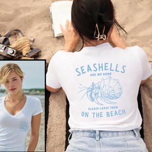 Op de afbeelding: Wit T-shirt met blauwe tekst en een schelpmotief. De tekst luidt "Seashells are my home, please leave them on the beach." De persoon draagt het T-shirt op een zandstrand, met een boek en een camera in de buurt.