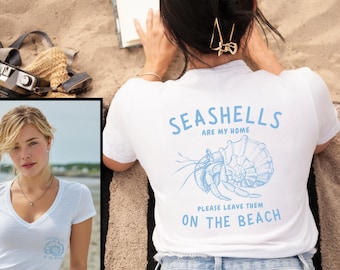 May include: White t-shirt with blue text and a seashell graphic. The text reads "Seashells are my home, please leave them on the beach." The shirt is worn by a person on a sandy beach, with a book and camera nearby.