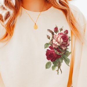 Op de afbeelding: Crèmekleurige sweatshirt met een bloemenontwerp met een slang die zich om rozen wikkelt. De rozen zijn dieprood en blushroze, met groene bladeren. Een gouden ketting met een hanger is ook zichtbaar.