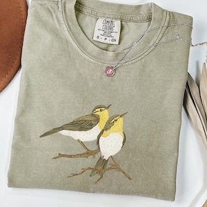 Op de afbeelding: Saliegroen t-shirt met een illustratie van twee geel-witte vogels op een tak. Een zilveren ketting met een roze hanger ligt op het shirt. Het label luidt "Comfort Colors".