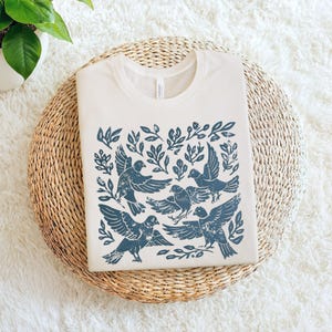 Pu&ograve; includere: T-shirt color crema con un design di uccelli e fogliame blu. La grafica include diversi uccelli in volo e appollaiati tra le foglie. La maglietta &egrave; piegata su un tappetino circolare intrecciato, con una pianta sullo sfondo.