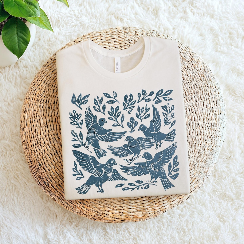 Pu&ograve; includere: T-shirt color crema con un design di uccelli e fogliame blu. La grafica include diversi uccelli in volo e appollaiati tra le foglie. La maglietta &egrave; piegata su un tappetino circolare intrecciato, con una pianta sullo sfondo.
