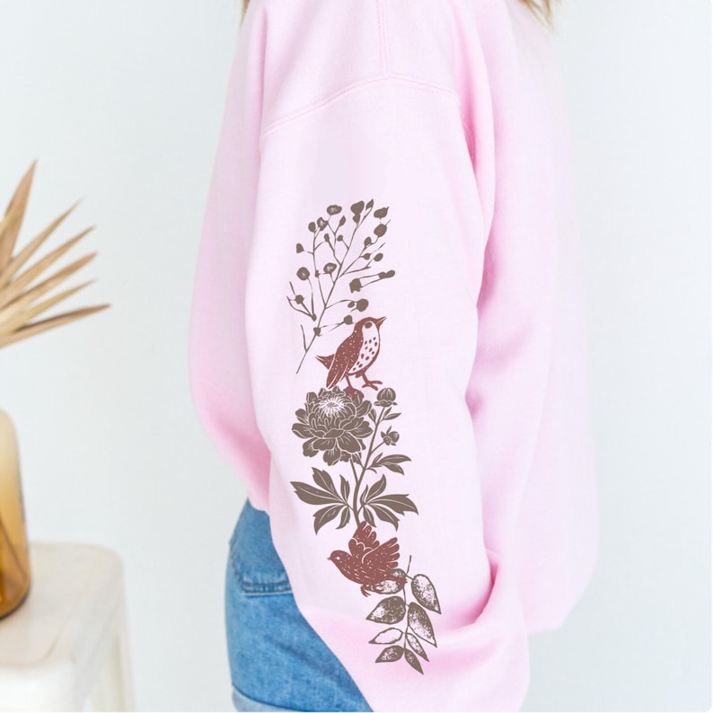 May include: A light pink sweatshirt with a brown floral and bird design on the left sleeve. The design includes a bird perched on a flower, leaves, and other floral elements. The sweatshirt is made of a soft material.