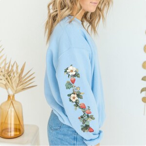 Peut inclure: Un sweat-shirt bleu clair avec un motif floral sur la manche. Le motif repr&eacute;sente des fleurs blanches et des fraises rouges.