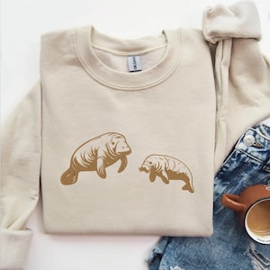 May include: A beige sweatshirt with a brown graphic of two manatees. The manatees are facing each other and the larger manatee is on the left side of the image.