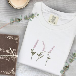 Op de afbeelding: Wit Comfort Colors T-shirt met drie geborduurde lavendeltakken. De takken hebben roze bloemen en groene stelen en bladeren. Een bruin leren notitieboek met decoratieve stiksels en een kaars staan ook in de afbeelding.