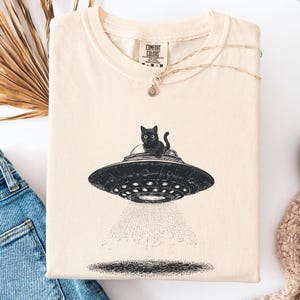 May include: Cream-colored t-shirt featuring a black graphic of a cat in a UFO. The UFO is emitting a beam of light. The shirt has a crew neck and short sleeves. The brand name "Comfort Colors" is visible on the tag.